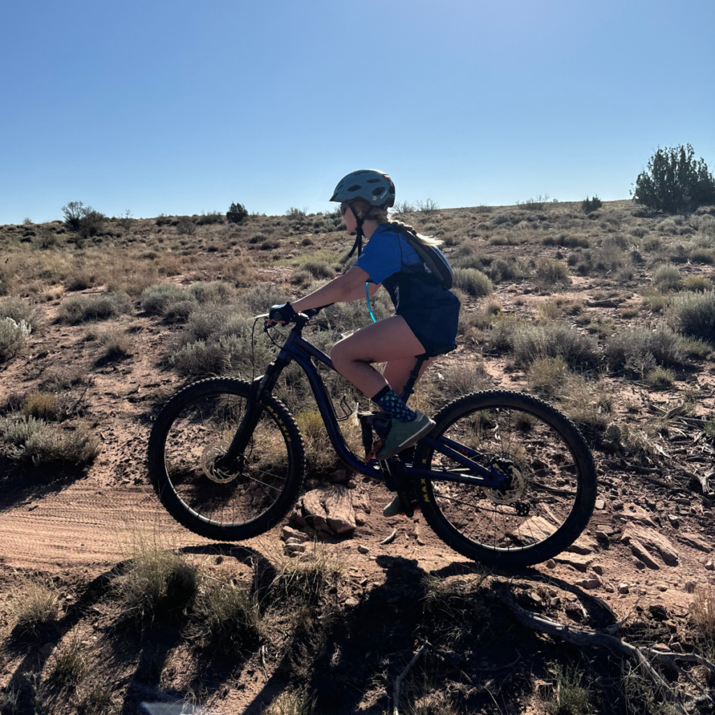 A young girl riding a mountain bike on a dirt trail, fully focused and determined, showcasing her love for the sport. The photo captures her in action during a race, highlighting both the thrill of competition and her connection to the outdoors.