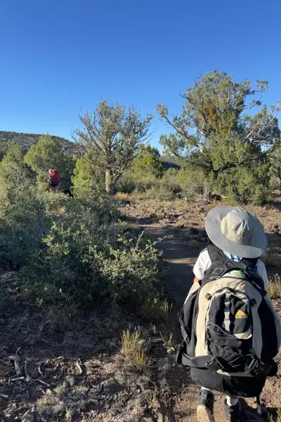 Young boy wearing a large backpack and wide-brim cooling hat, walking on a desert trail under a bright blue sky, with another hiker in the distance.